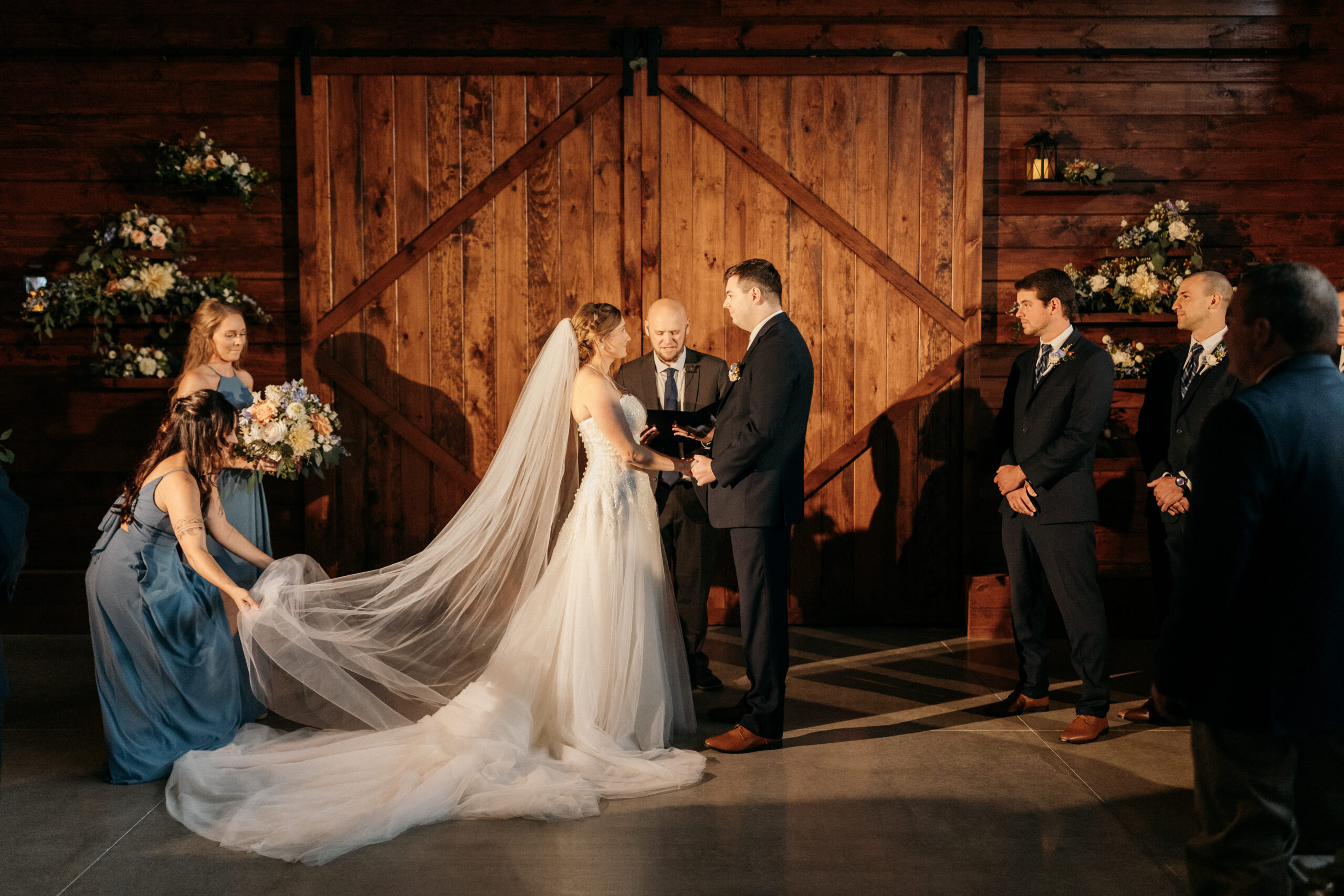 Bride and Groom saying I do at the alter in a rustic barn setting. The maid of honor holding up the bride's veil and the bride and groom holding hands, looking into each others eyes.