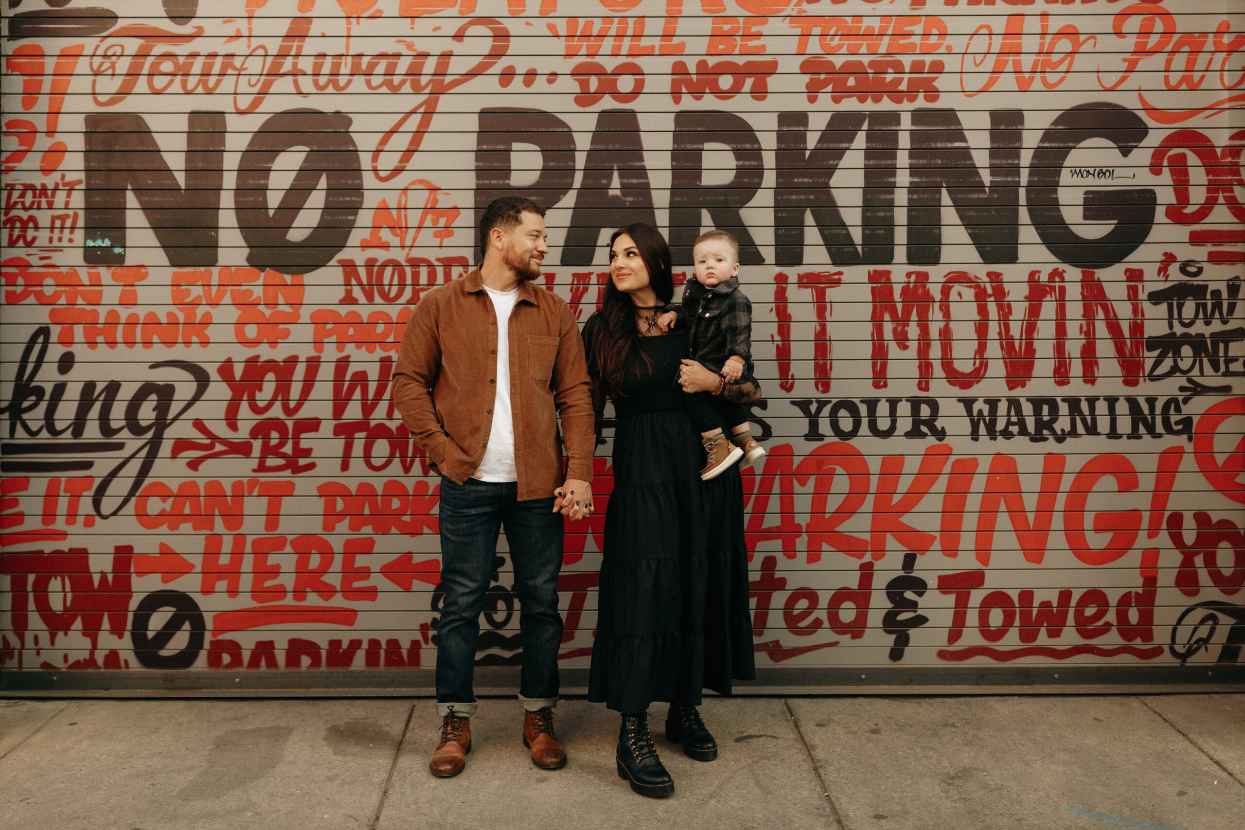 Standing infront of a brick wall filled with red and black spray painted words. Madison is holding their son Jackson, with Tim standing on her other side looking down at her and his son.