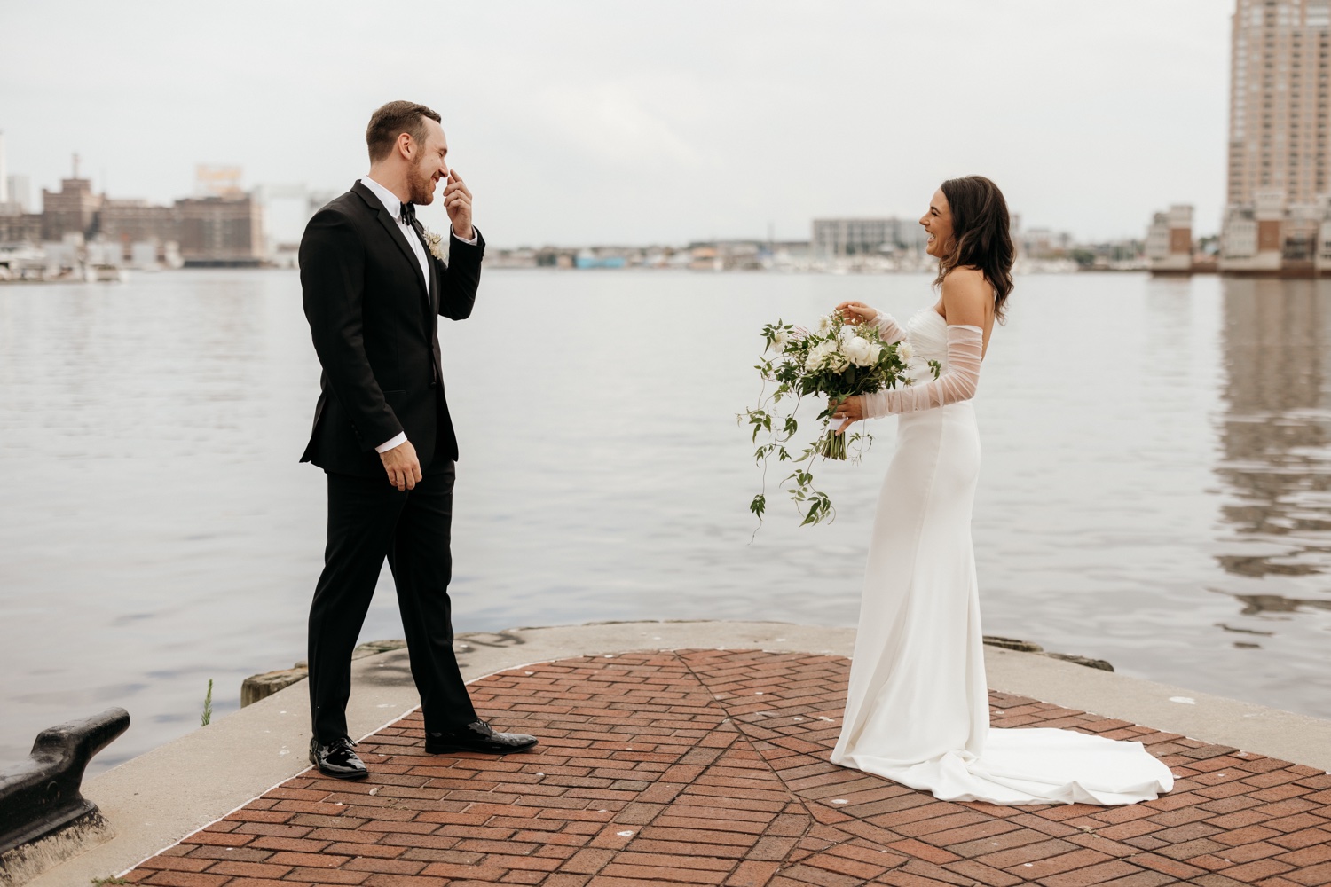 A bride and groom having their first look by the water. The bride is laughing and smiling while the groom is covering his face in shock and happiness.