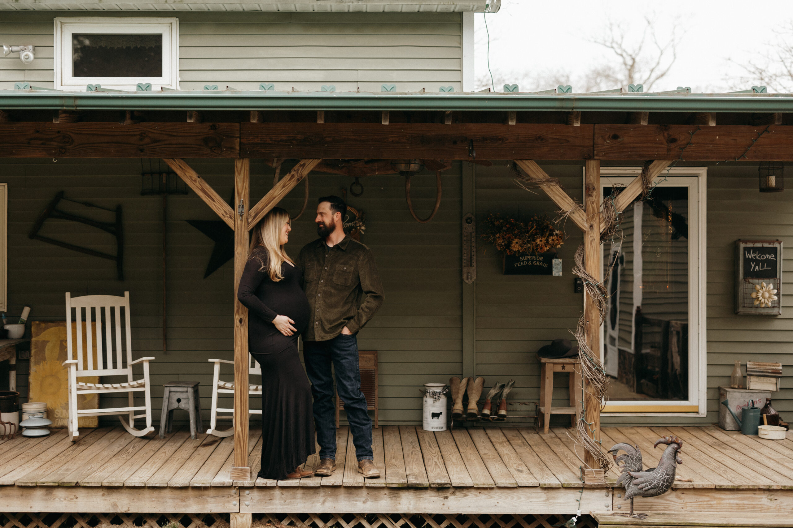 Maternity Session at a Farm house. The couple is standing on their porch. Mom is up against a post wearing a long black dress and dad is standing beside her looking down at her face.