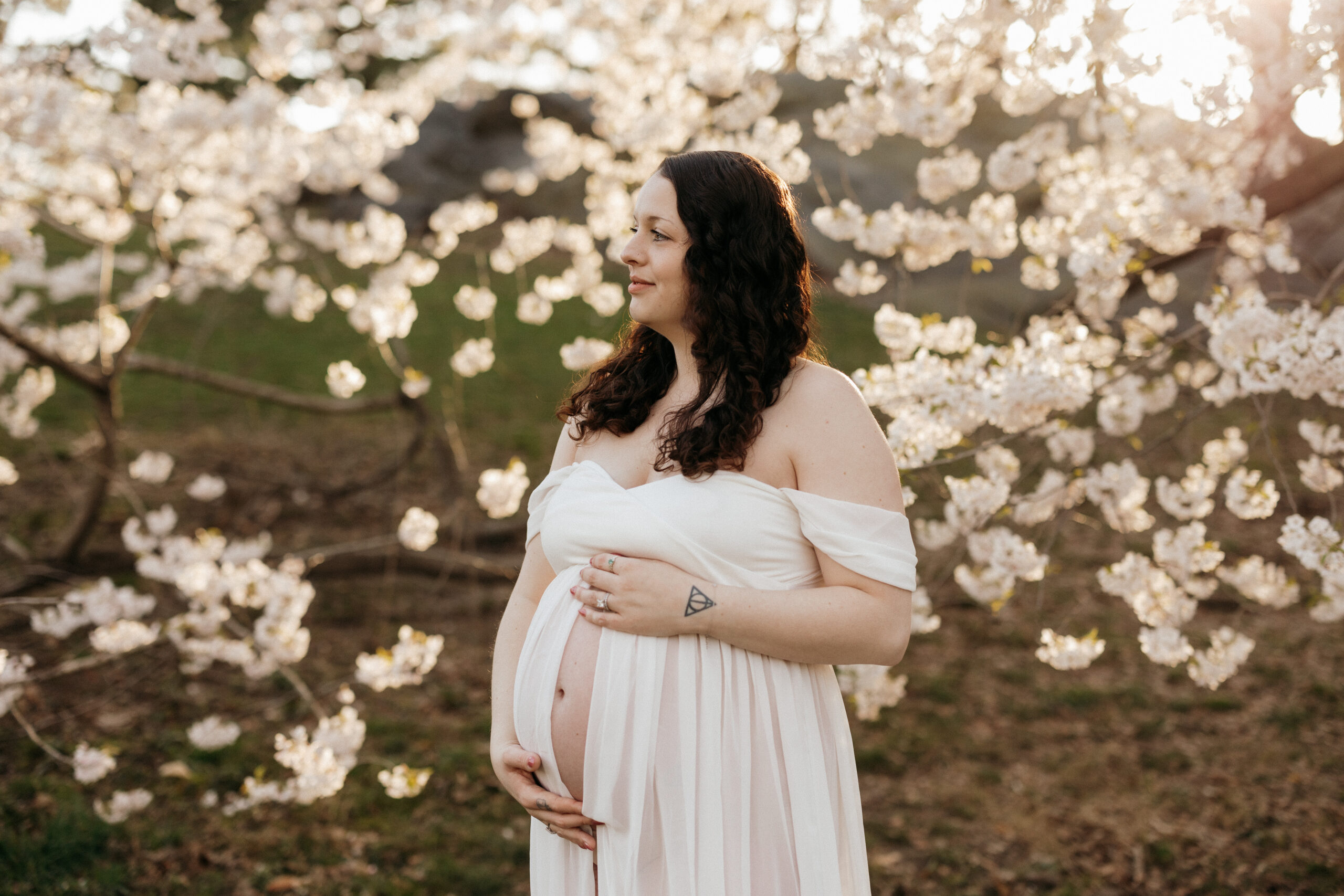 Pregnant mom standing in front of a cherry blossom tree, sun light shining in between the branches. Mom is wearing a flowing white dress, has her hands on her pregnant belly, and looking out into the park.