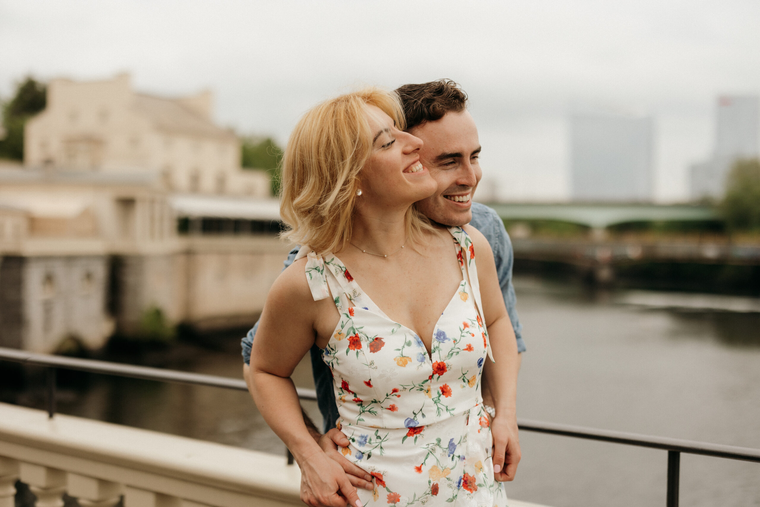 A couple standing on a bridge. The male figure behind the female, wrapping his hands around her as they look into the distance smiling. The unfocused water background keeps them focused in the center of the photo.