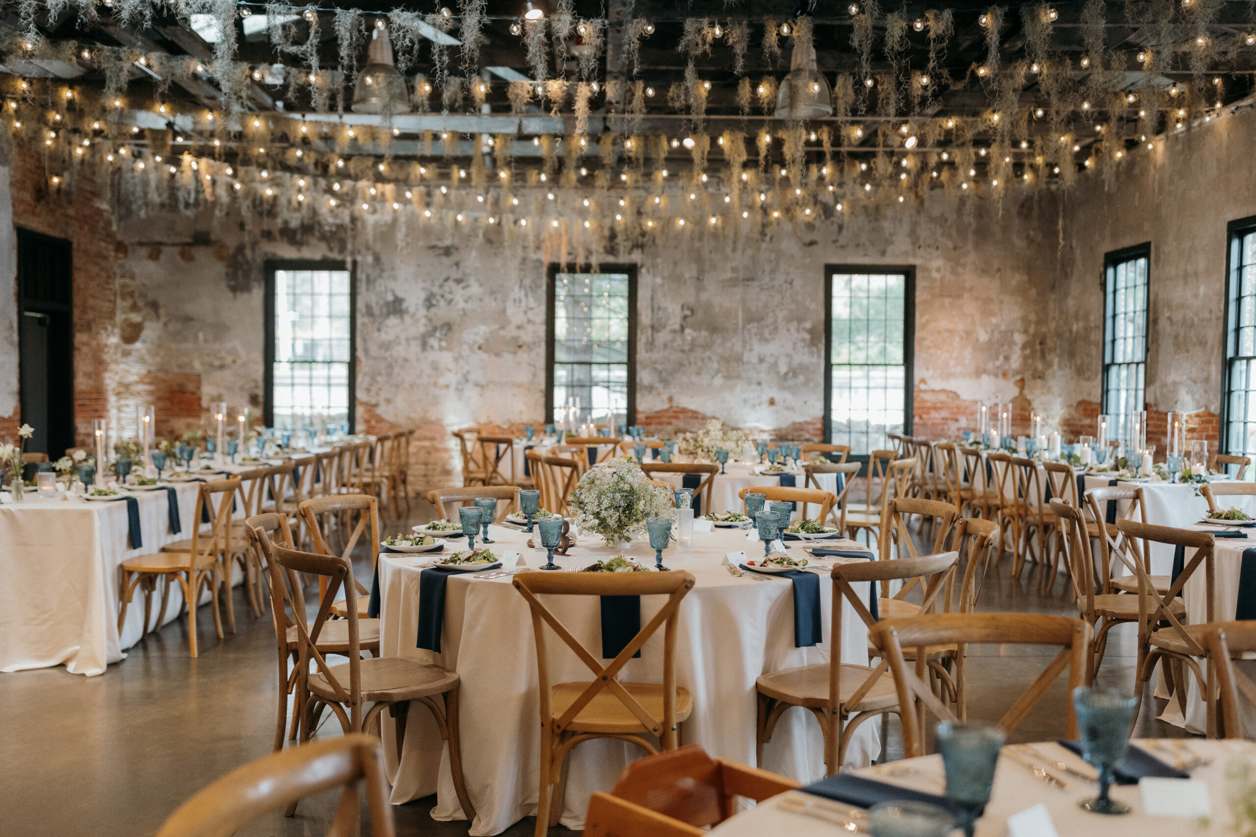 A decorated room for a wedding with hanging little lights, a large brick wall at the back. Brown wooden timeless chairs, beige, blue, and white colour scheme. Large blue glasses on each table with beautiful floral piece sin the center.