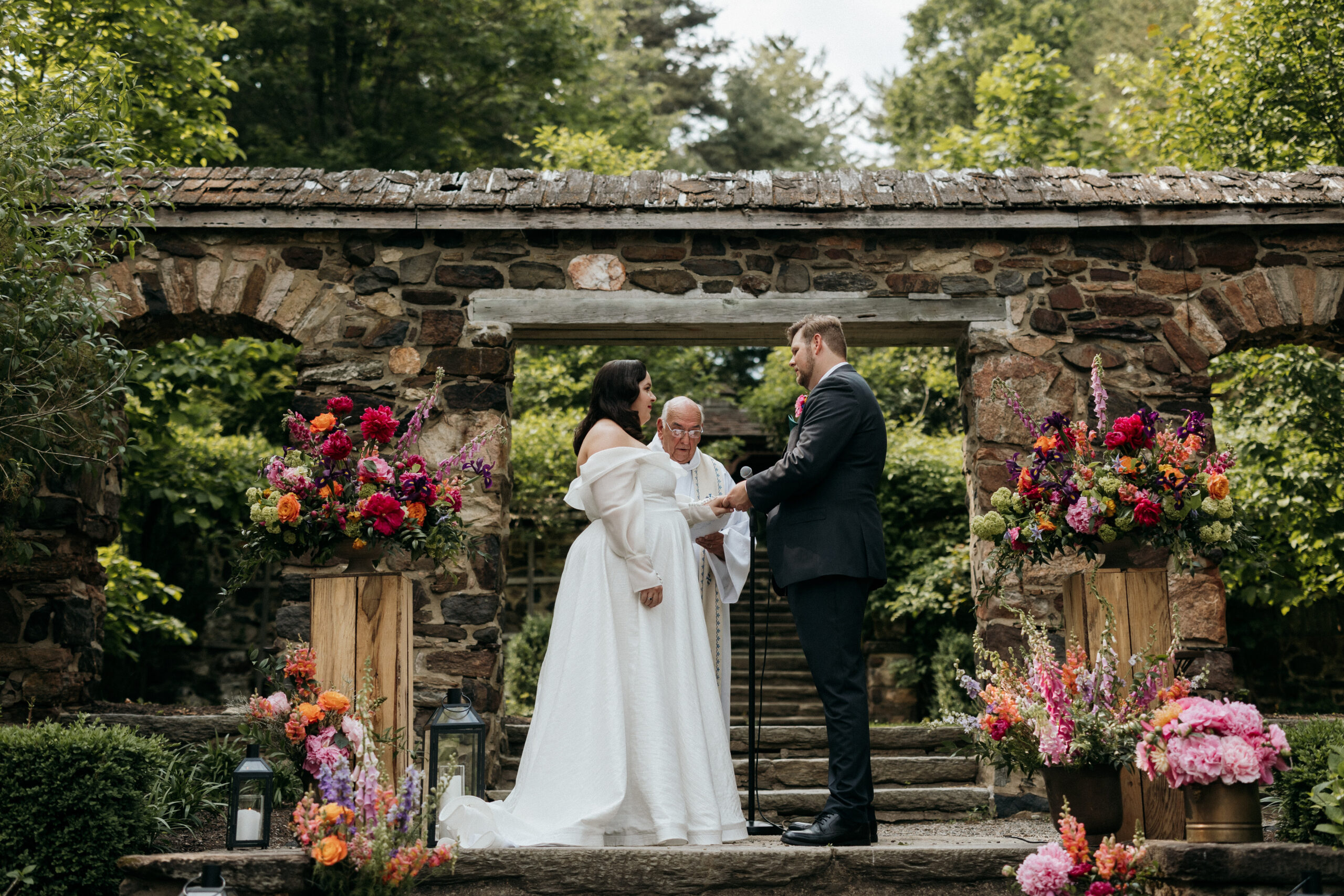 Up on a very beautiful rustic staircase in the middle of a forest, the bride and groom are looking into one another eyes getting married. Two beautoful flower arrangements are on either side on them, the priest in between them up on the stairs.