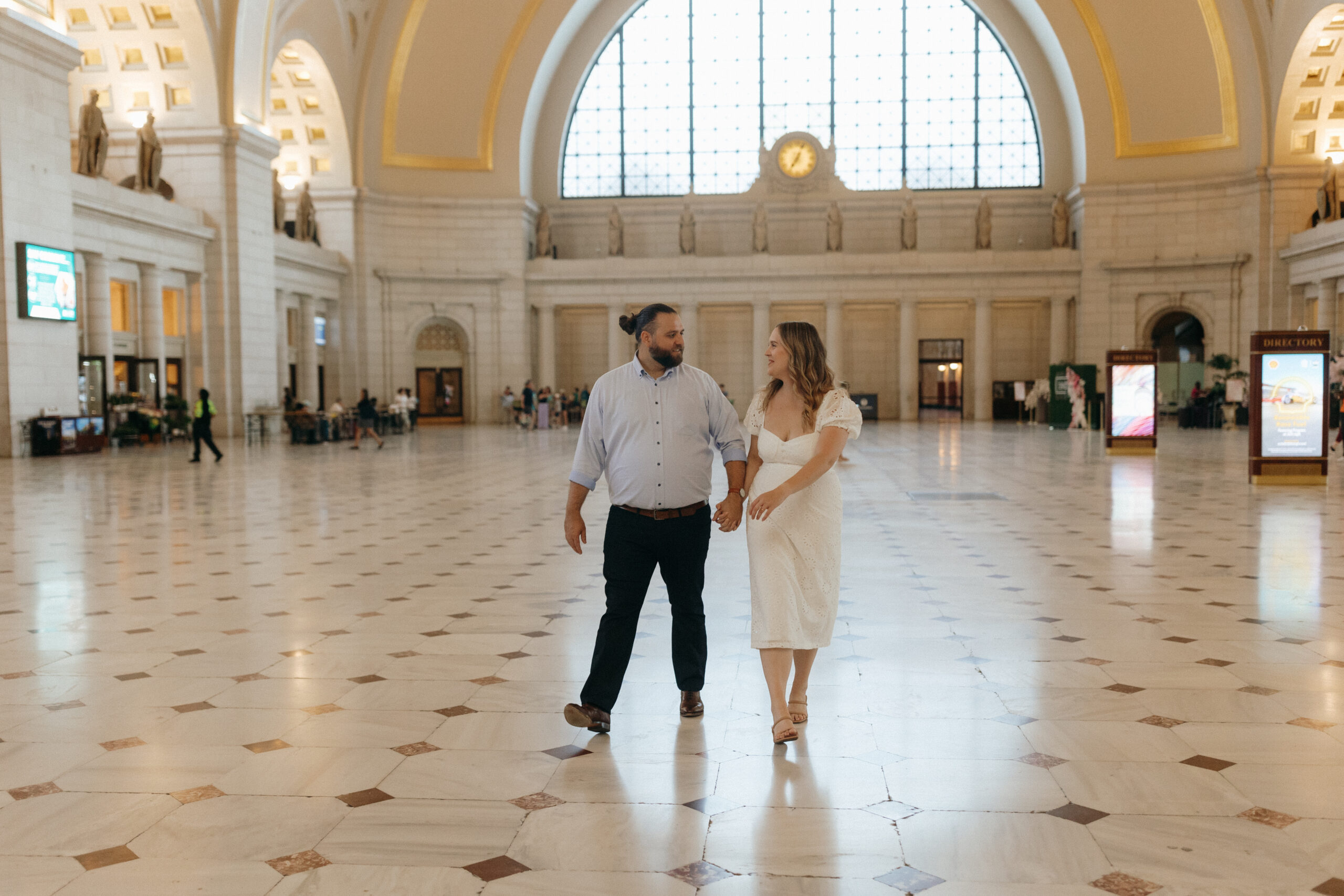 Meghan (wearing a flowy white dress and has curled blonde hair) and Tyler (wearing a dress shit and dress pants with dark brown hair tied in a bun) walking hand in hand through union station in Washington, DC.