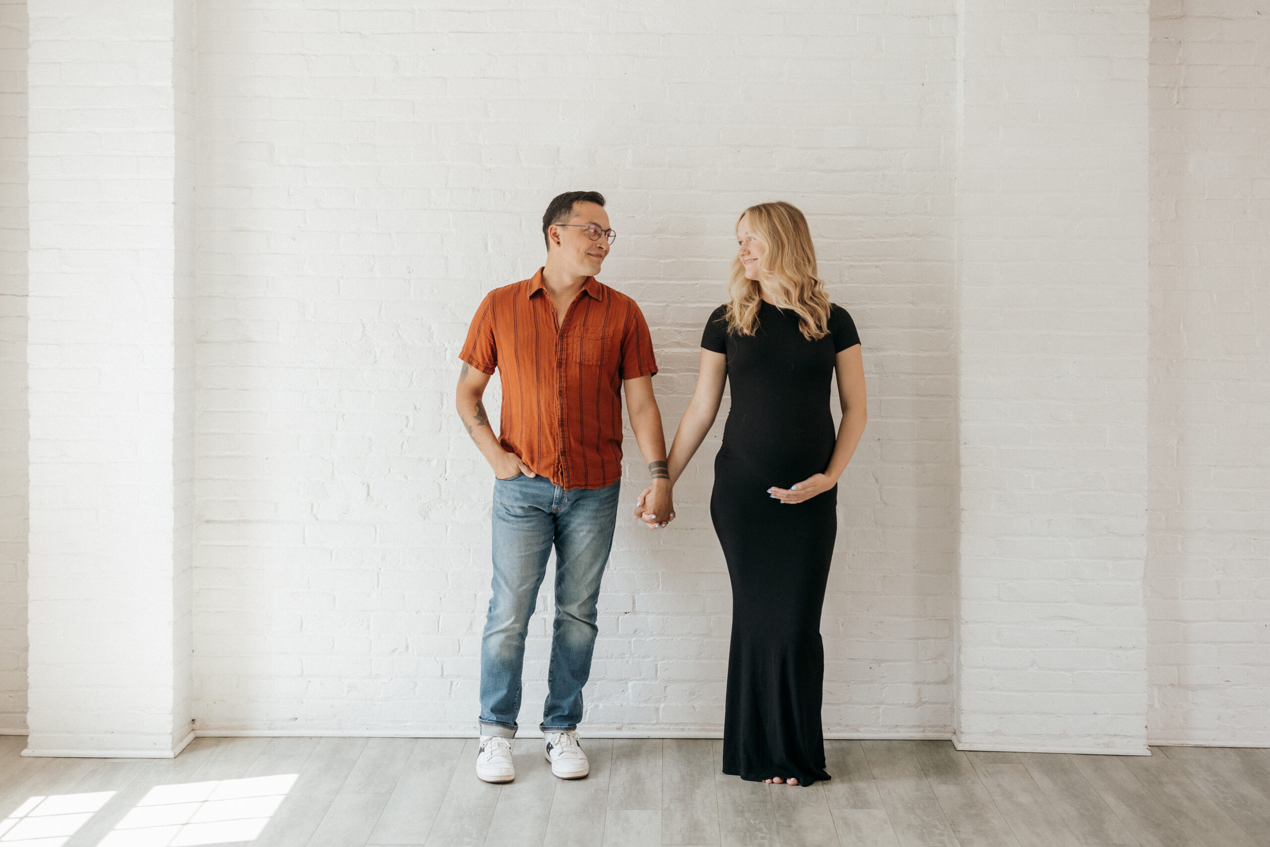 The couple is leaning against a white bricked wall facing the camera. Ruben on the left, wearing light blue jeans, and orangy red button up t-shirt and white sneakers and Emily on the right, blonde hair (curled) and wearing a long black t-shirt dress. They are both holding hands, emily has her left had on her belly, and looking at one another smiling.m