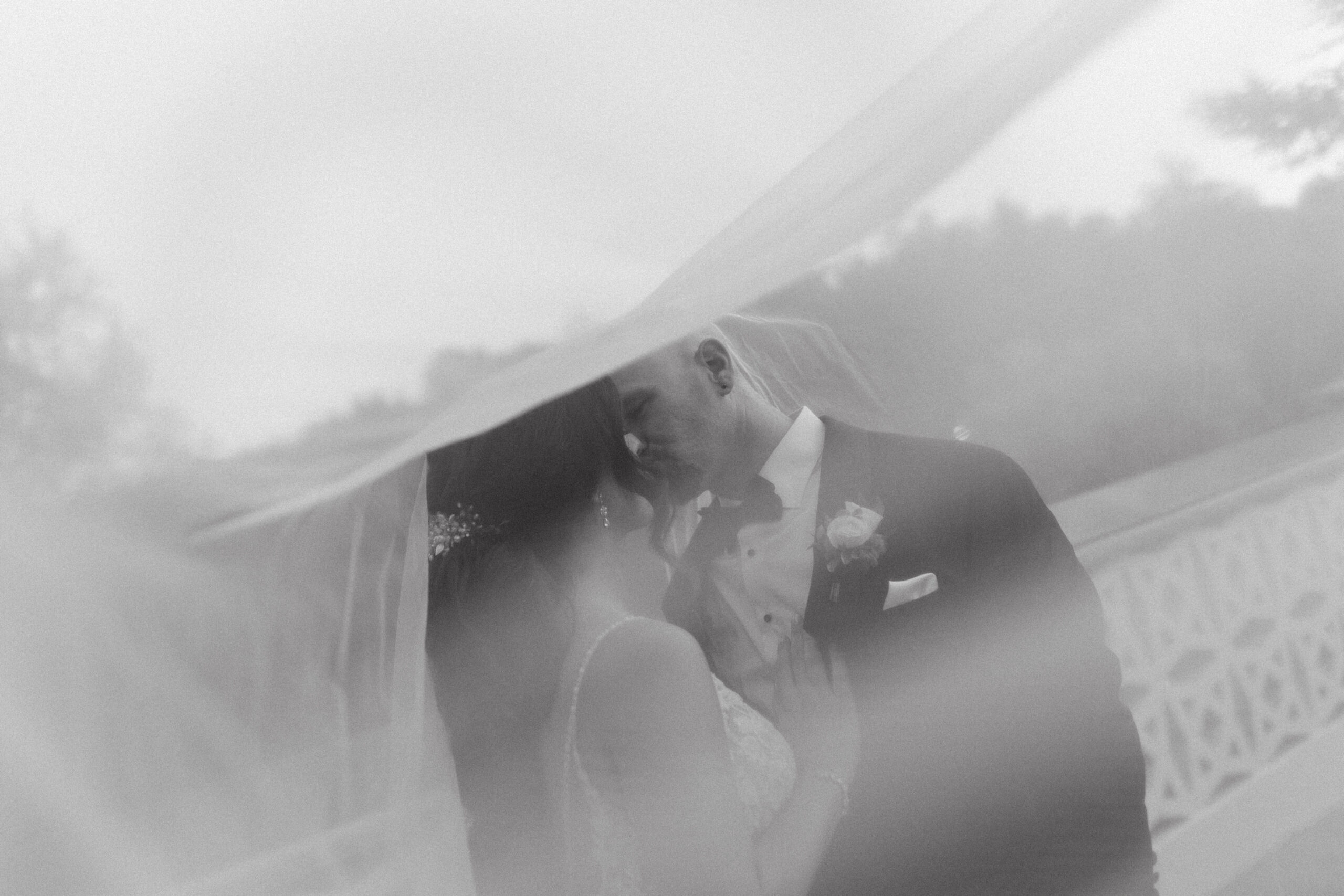 A black and white photo of the brides's veil blowing in the wind towards the camera. Bride and Groom are embracing in a kiss.