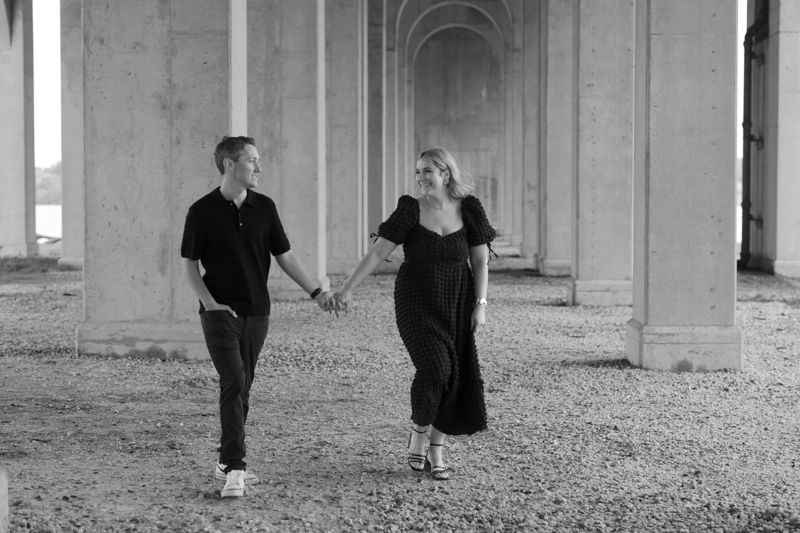 Couple is walking through on gravel stones underneath a concrete arches. Both looking at each other smiling hand in hand.